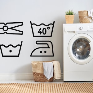 Op de afbeelding: Zwart-witte wassymbolen, waaronder een wasknijper, wassen op 40 graden, niet bleken en strijken. Een witte wasmachine, een geweven mand met wasgoed en decoratieve items staan op de achtergrond.