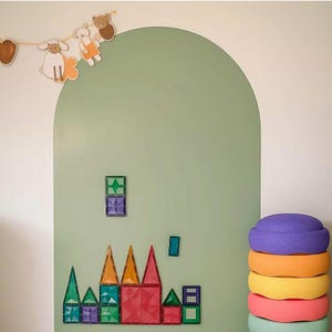 Op de afbeelding: Een saliegroene boogvormige muursticker met een slinger van papieren dierenfiguren bovenaan. Magnetische bouwtegels in diverse geometrische vormen en kleuren zijn op het oppervlak geplaatst, naast een stapel van vijf kleurrijke balanceerstenen.