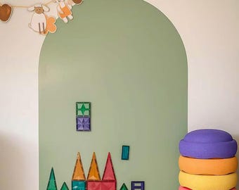May include: A sage green arched wall decal featuring a string of animal-themed paper cutouts at the top. Magnetic building tiles in various geometric shapes and colors are arranged on the wall. A stack of five colorful, rounded foam balance stones.