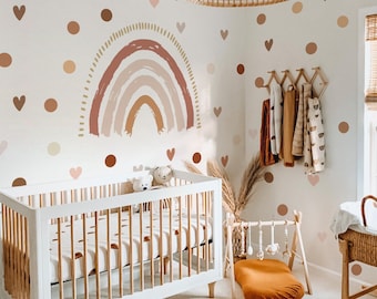 May include: A nursery with a white crib, a wooden play gym, and a woven light fixture. The walls are decorated with a rainbow, hearts, and polka dots in shades of brown, pink, and beige. A wooden rack holds several blankets and clothing items.