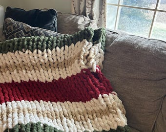 May include: A chunky knit blanket in a color block design of green, white, red, and tan. The blanket is draped over a gray couch, with pillows visible in the background. The thick yarn creates a cozy, textured appearance.