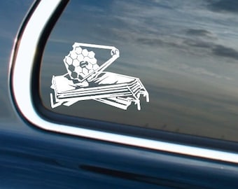 May include: A white vinyl decal of the James Webb Space Telescope, affixed to the rear window of a dark-colored vehicle. The decal shows the telescope's distinctive hexagonal mirror and sunshield, with a detailed design. The background shows a cloudy sky.