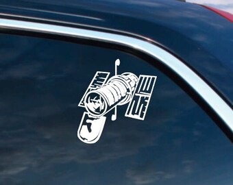 May include: White vinyl decal of the Hubble Space Telescope on a car window. The decal shows the telescope's cylindrical body, solar panels, and other components. The background shows a cloudy sky and the reflection of the sun.