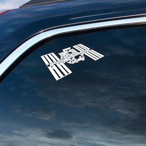 May include: White vinyl decal of the International Space Station (ISS) on a car window. The decal is a detailed outline of the ISS, with solar panels and modules visible. The car is black, and the window reflects a cloudy sky.