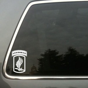 May include: White vinyl decal featuring the word "AIRBORNE" above a shield design with a winged blade. The decal is affixed to a car window, with a blurred outdoor scene visible through the glass. The design is a military emblem.