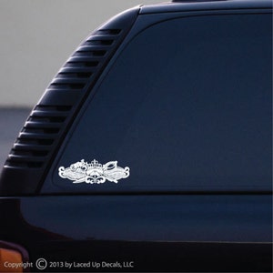 May include: A silver decal featuring a detailed emblem with a crown, wings, and other nautical elements. The decal is affixed to a dark vehicle window, showcasing its intricate design and reflective surface.
