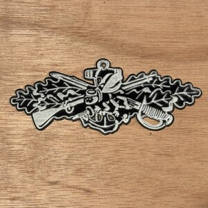 May include: Black and white embroidered patch featuring a detailed emblem with a rifle, sword, anchor, and oak leaves. The patch has a white border and is likely a military or nautical insignia.