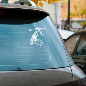 Può includere: Adesivo in vinile bianco raffigurante un satellite su un finestrino dell'auto. L'adesivo presenta un'illustrazione dettagliata di un satellite con pannelli solari e antenne. L'auto è marrone scuro, con uno sfondo sfocato di alberi ed edifici.