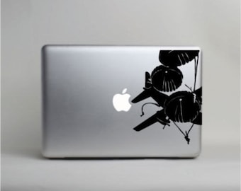 Puede incluir: Un portátil plateado con una calcomanía negra de paracaídas y aviones estilizados en la esquina. El logotipo de Apple es visible. Copyright 2013 by Laced Up Decals, LLC está impreso en la parte inferior.