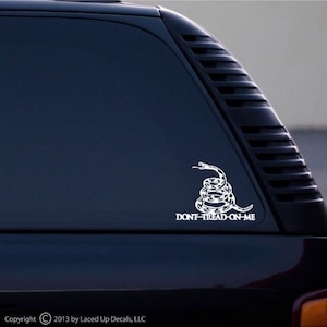 May include: White vinyl decal featuring a coiled rattlesnake above the text "DON'T TREAD ON ME." The decal is applied to a vehicle window. The design is a symbol of American libertarianism and individual rights.