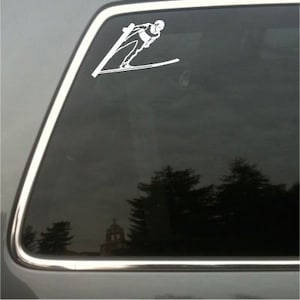 May include: White vinyl decal of a ski jumper in mid-air, affixed to a car window. The skier is depicted in a dynamic pose, with skis extended. The background shows a reflection of trees and a building.