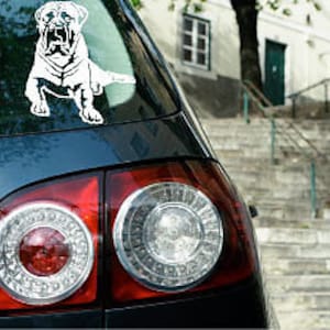 Puede incluir: Una calcomanía de vinilo blanco de un perro, adherida a la ventana trasera de un coche oscuro. El perro, posiblemente un mastín, está en posición sentada. Se ven los faros traseros del coche.