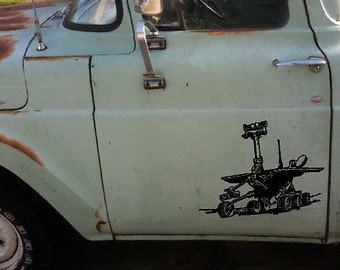 May include: A black decal of a Mars rover is affixed to the side of a vintage, light green pickup truck. The truck shows signs of age with rust and wear. The rover design is detailed, featuring wheels, a central mast, and various scientific instruments.