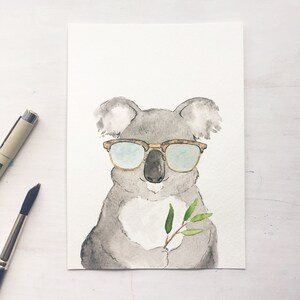 May include: Watercolor painting of a gray koala wearing brown-rimmed sunglasses. The koala holds a sprig of green leaves. A black pen and paintbrush are in the background. The artwork is on a white background.