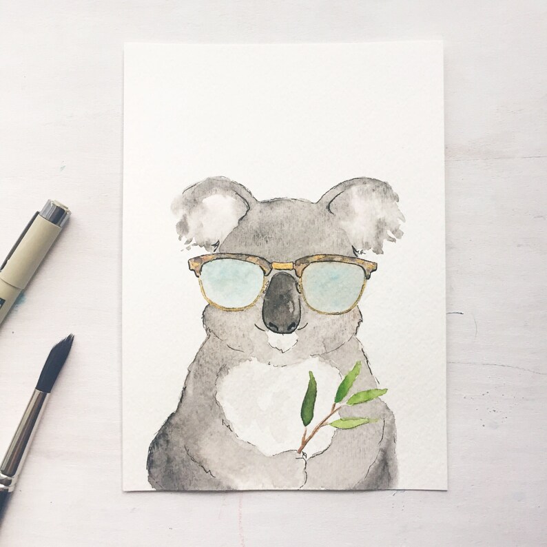 May include: Watercolor painting of a gray koala wearing brown-rimmed sunglasses. The koala holds a sprig of green leaves. A black pen and paintbrush are in the background. The artwork is on a white background.