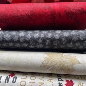 May include: A stack of fabric rolls featuring Canadian-themed designs. The fabrics display maple leaves in red, black, and gold, along with text that includes the words "Our Home and Native Land". The top fabric has a red and white pattern.