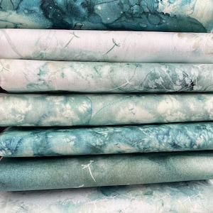 May include: A stack of folded fabric bolts featuring various shades of teal, turquoise, and white. The patterns include abstract watercolour washes, botanical silhouettes, and delicate dandelion seed motifs printed across the textured textile surfaces.