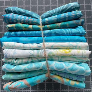 Peut inclure: Une pile de dix carrés de tissu bleu et blanc à motifs, liés ensemble avec de la ficelle. Le tissu a un motif batik.