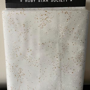 Puede incluir: Un rollo de tela con fondo gris claro y un patrón de motas blancas y doradas. El borde superior presenta una banda negra con el texto "RUBY STAR SOCIETY". Esta tela es adecuada para proyectos de acolchado y costura.