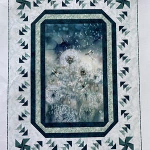 Op de afbeelding: Een quilt met een centraal beeld van paardenbloemen in aquarel. De quilt heeft een donkerblauwe en lichtgroene rand met windmolen- en driehoekaccenten. Het kleurenschema is zacht en gedempt.