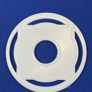May include: A white, circular object with a central hole and an outer ring. The design includes four square-shaped cutouts around the central hole. The object is made of a textured material and is set against a blue background.