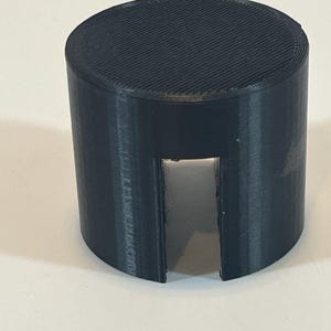 May include: A cylindrical, black object with a slot cut out of the side. The top surface has a ridged texture. The object appears to be made of plastic and is likely a component or part of a larger item.