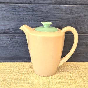 May include: A light peach-coloured ceramic teapot with a smooth, matte finish. The teapot features a curved handle, a pointed spout, and a removable lid topped with a small, circular, light mint green knob that contrasts with the peach body.