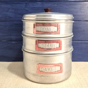 May include: A three-tiered, vintage metal canister set with a brown knob on top. Each section is labelled with a pink and white label: Scones, Biscuits, and Cakes. The canisters are silver with a ridged design.