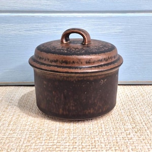 May include: A brown ceramic jar with a lid and handle. The jar is round and has a textured surface. The lid has a curved handle. The jar is on a beige surface.