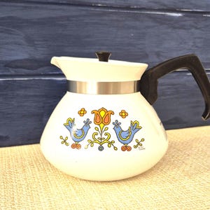 May include: A white vintage coffee pot with a black handle and lid. The pot has a decorative design of two blue birds, orange and yellow flowers, and a silver band. The background is a dark blue wooden surface.