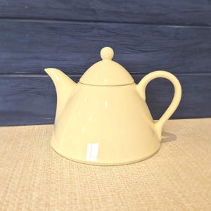 May include: A cream-coloured ceramic teapot with a smooth, glossy finish. The teapot features a wide, rounded base, a curved spout, a C-shaped handle, and a matching lid topped with a small, spherical knob. The design is simple and functional.