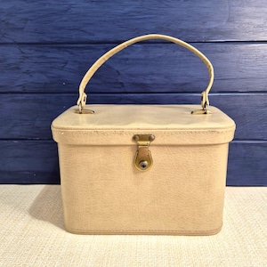 May include: A vintage beige rectangular vanity case with a curved handle and a gold-coloured clasp. The small travel case is made of a textured material and sits on a light-coloured surface against a blue background.