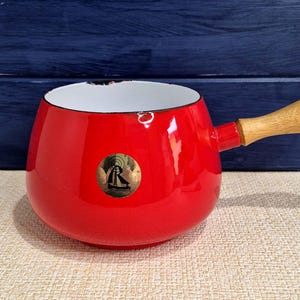 May include: A bright red enamel saucepan with a wooden handle. The small pot has a white interior and a gold emblem on the side. The handle is made of light-coloured wood. A charming kitchenware item.