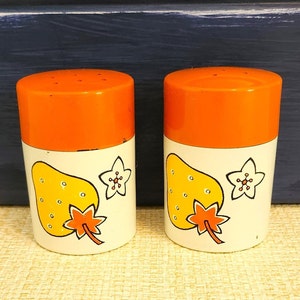 May include: A pair of orange and white ceramic salt and pepper shakers featuring a strawberry design. The shakers have a white background with a yellow strawberry, a green leaf, and a white flower.
