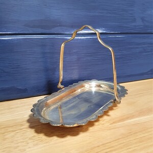May include: A silver-coloured, oval serving tray with a scalloped edge and a handle. The handle is arched and attached to the tray's sides. The tray is set on a wooden surface with a blue background.