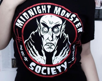May include: A black t-shirt with a red and white circular graphic of a vampire with the text "Midnight Monster Society".