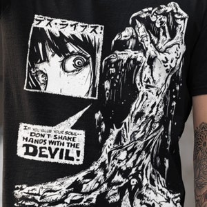 May include: Black and white graphic t-shirt with a comic book style illustration of a monstrous hand reaching out from a dark background. The text "デス・ライブズ!" is in the top left corner and the text "IF YOU VALUE YOUR SOUL-- DON'T SHAKE HANDS WITH THE DEVIL!" is in the bottom left corner.