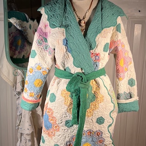 以下が含まれることがあります： パステルカラーの花柄のキルトローブ。緑色のショールカラーと、お揃いのリボンベルトが特徴です。マネキンに着せられており、パッチワークのデザインとスカラップの縁が際立っています。