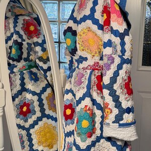 Puede incluir: Una chaqueta acolchada azul y blanca con capucha, con un diseño de patchwork de flores de colores. La chaqueta se lleva sobre una falda azul.