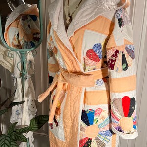 Puede incluir: Una bata acolchada de color melocotón con capucha blanca y diseño de patchwork. La bata tiene un cinturón de atar y está hecha de una tela suave y cómoda.