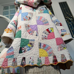 Puede incluir: Una colcha blanca con capucha, borde festoneado y diseño de patchwork de retazos de tela de colores. La colcha está formada por cuadrados con un patrón en forma de abanico en cada cuadrado.