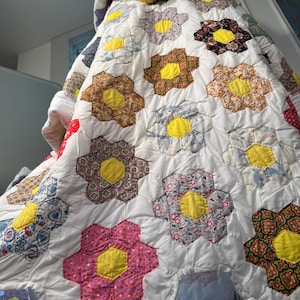 Pode incluir: Um edredom vintage com um padrão floral repetitivo. O edredom apresenta grandes flores de seis pétalas em várias cores, incluindo rosa, castanho e azul, cada uma com um centro amarelo. As flores estão sobre um fundo branco.