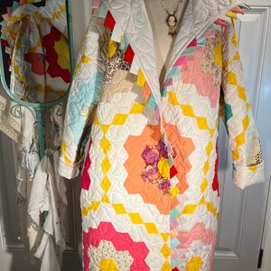 May include: A white quilted coat with a colourful patchwork design. The coat has a large collar and a long, flowing silhouette. The patchwork design features a variety of floral patterns in shades of pink, yellow, and blue.