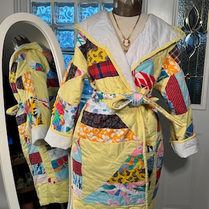 Op de afbeelding: Een gele gewatteerde badjas met een patchwork-ontwerp met verschillende kleuren en patronen, waaronder rode, blauwe, groene en bloemenprints. De badjas heeft een capuchon en een strikceintuur.