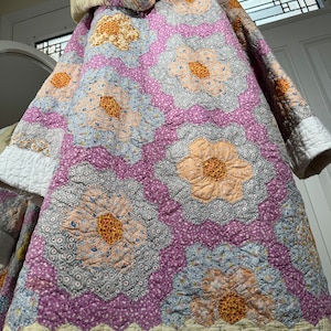 May include: A purple and white quilt with a floral pattern. The quilt is made of patchwork fabric and has a hood.