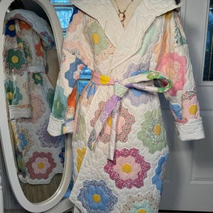 Puede incluir: Una bata blanca acolchada con un patrón floral. La bata tiene una capucha y un cinturón de lazo. La bata está hecha de tela de patchwork con una variedad de colores, incluyendo rosa, azul, verde y amarillo.