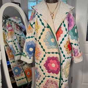 Könnte beinhalten: Ein weißer gesteppter Mantel mit einem grün-weißen Blumenmuster. Der Mantel hat eine Kapuze und lange Ärmel. Der Mantel ist aus Patchwork-Stoff gefertigt und hat einen Vintage-Look.