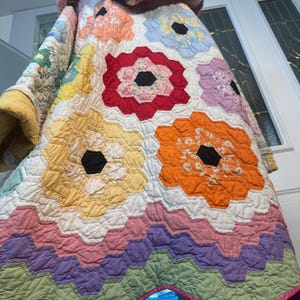 Könnte beinhalten: Ein farbenfrohes, handgefertigtes Steppdecke mit großen Blumenmustern in Rot-, Orange-, Gelb- und Blautönen. Die Steppdecke hat einen weißen Hintergrund und einen gewellten Rand mit rosa Besatz.