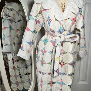 Puede incluir: Una bata blanca acolchada con cuello festoneado y cinturón de lazo. La bata está hecha de tela de patchwork con una variedad de colores y patrones. La bata se muestra en un maniquí con un collar.