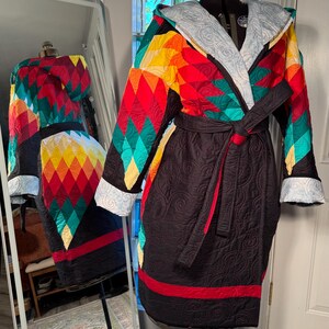 Puede incluir: Bata acolchada con un colorido estampado geométrico en la parte superior y las mangas. La bata tiene una base negra con una franja roja cerca del dobladillo y un cinturón negro. La capucha y los puños tienen un forro azul claro.